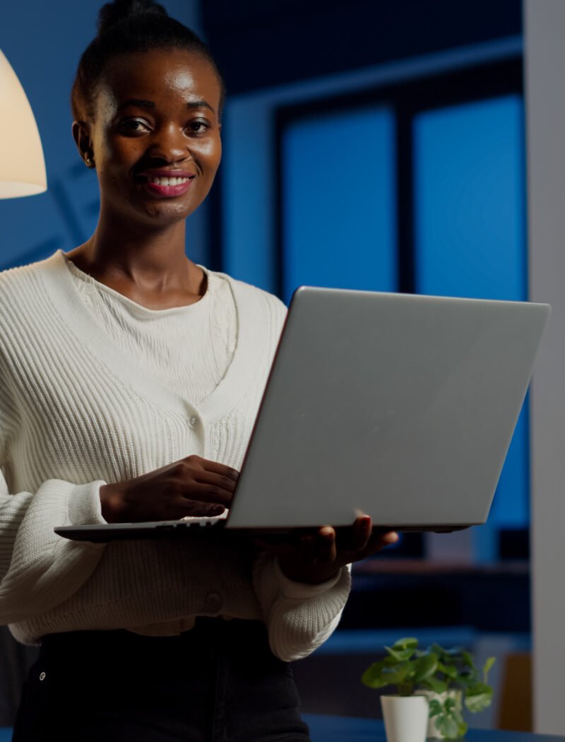 Black business woman looking at camera smiling holding laptop standing near desk in start-up company late at night. Focused employee using technology network wireless doing overtime for job